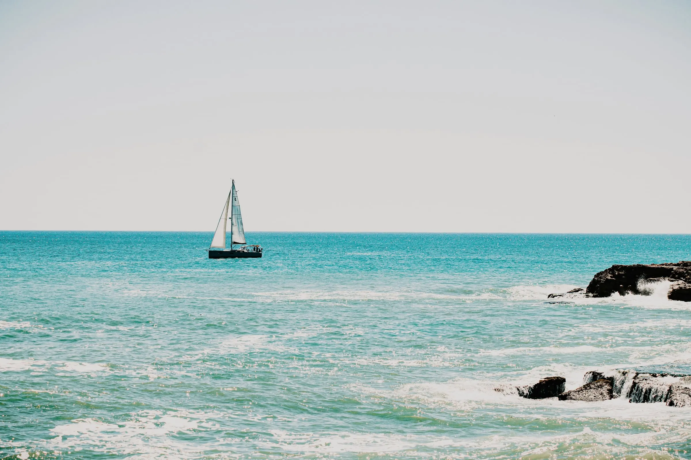 A sailboat on turquoise water near dark rocks along the shore