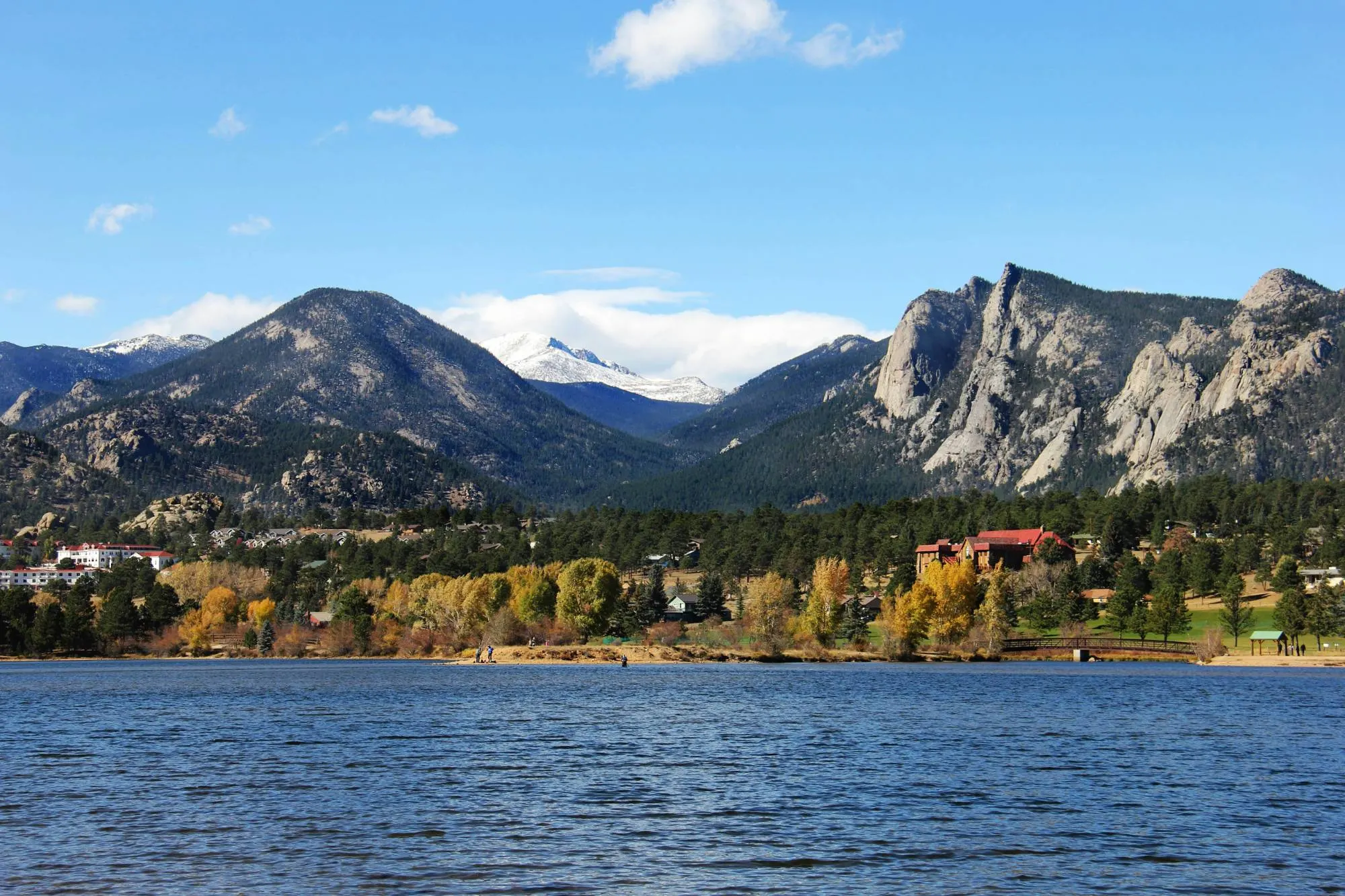 A mountain lake lined with autumn trees and homes beneath rocky, snow-dusted peaks